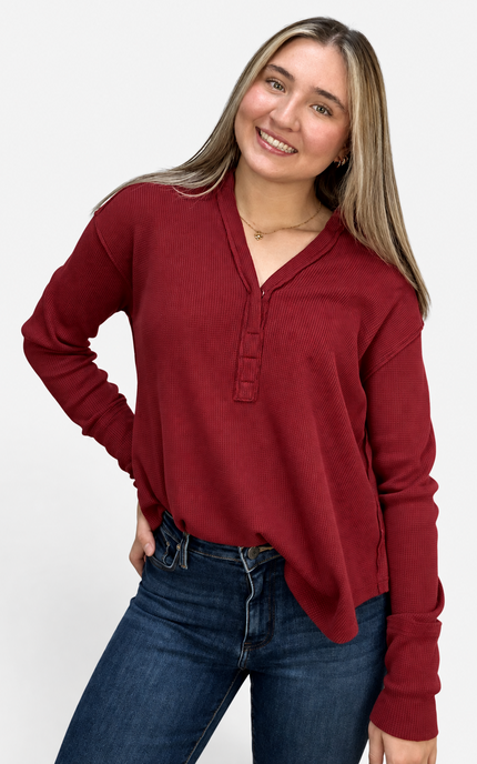 A woman wearing a long-sleeve henley-style top in deep burgundy red, paired with dark blue jeans, posing against a light gray studio background.