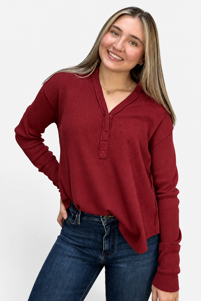 A woman wearing a long-sleeve henley-style top in deep burgundy red, paired with dark blue jeans, posing against a light gray studio background.