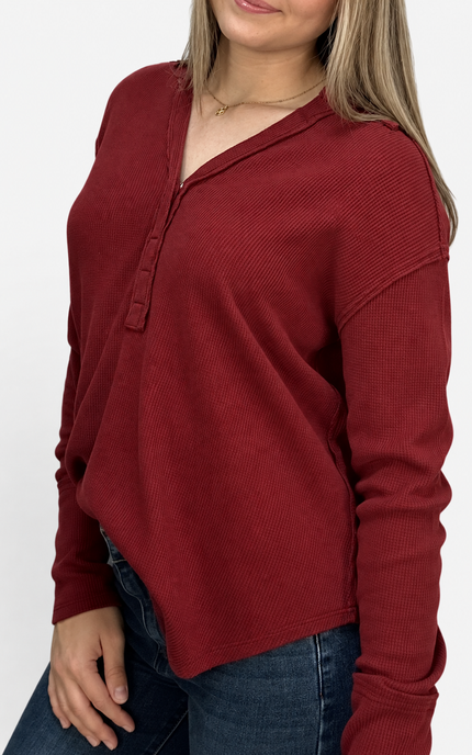 A woman wearing a ribbed long-sleeve henley top in deep burgundy red, paired with blue jeans, shown from the chest down against a plain white background.