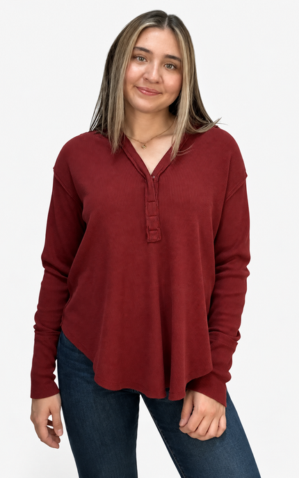A woman modeling a long-sleeve henley top in a deep burgundy red color, paired with dark blue jeans, against a plain white studio background.