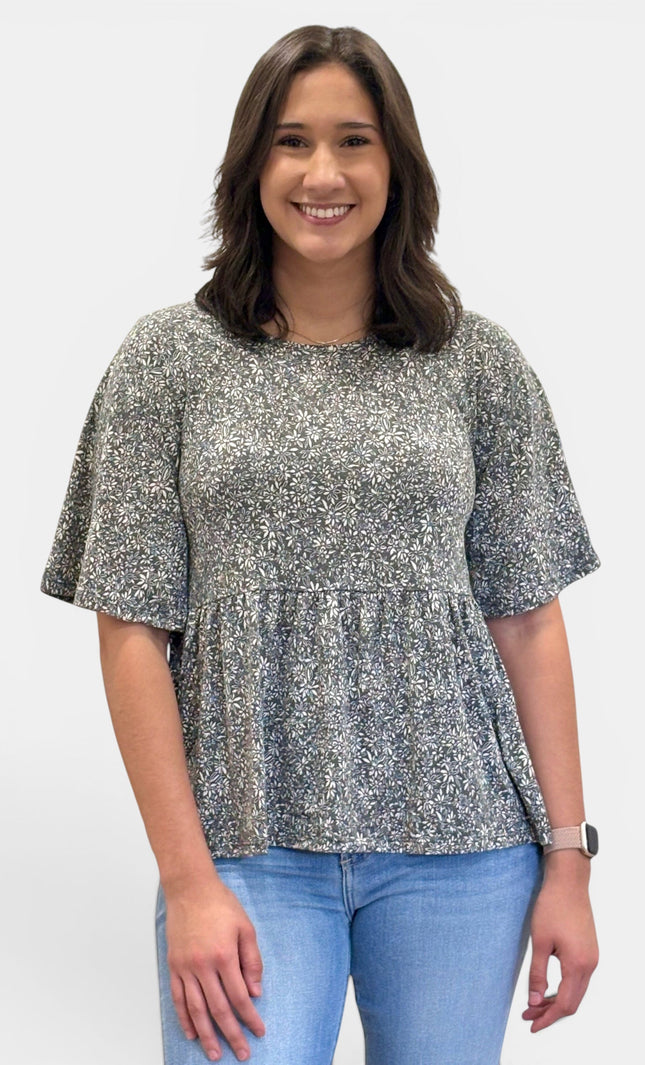 A woman wearing a short-sleeve peplum top with a gray and white floral pattern, paired with light blue jeans, standing against a plain light gray background.