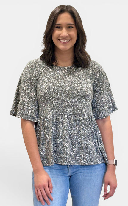 A woman wearing a short-sleeve peplum top with a gray and white floral pattern, paired with light blue jeans, standing against a plain light gray background.