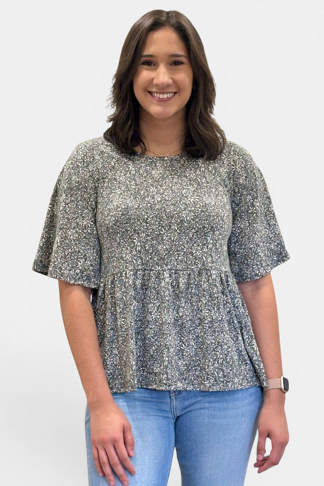 A woman wearing a short-sleeve peplum top with a gray and white floral pattern, paired with light blue jeans, standing against a plain light gray background.