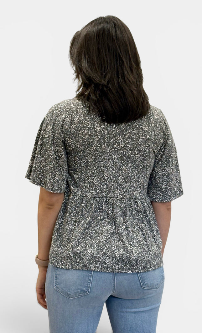 A woman viewed from the back wearing a short-sleeve peplum top with a black and white floral pattern, paired with light blue jeans, against a plain light gray background.