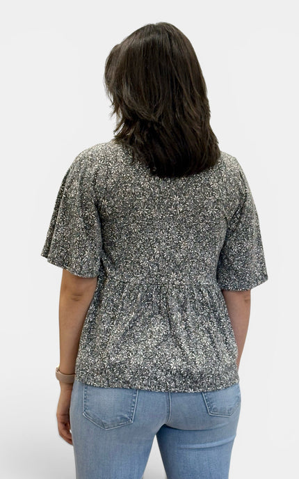 A woman viewed from the back wearing a short-sleeve peplum top with a black and white floral pattern, paired with light blue jeans, against a plain light gray background.
