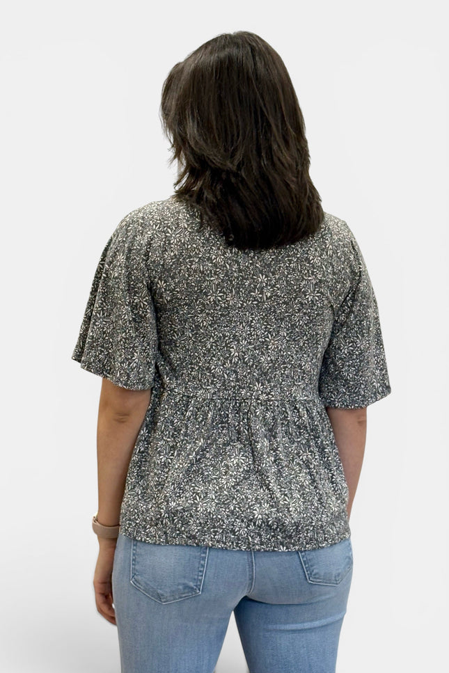 A woman viewed from the back wearing a short-sleeve peplum top with a black and white floral pattern, paired with light blue jeans, against a plain light gray background.
