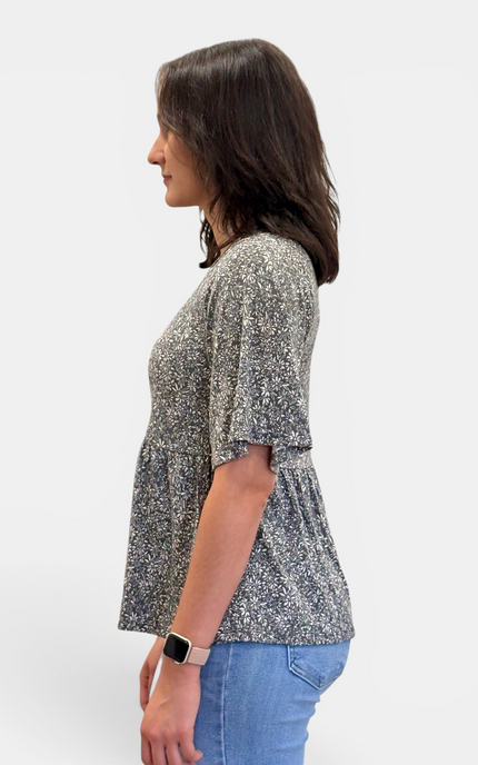 A woman shown in side profile wearing a short-sleeve peplum top with a gray and white floral pattern, paired with blue jeans, against a plain light gray background.