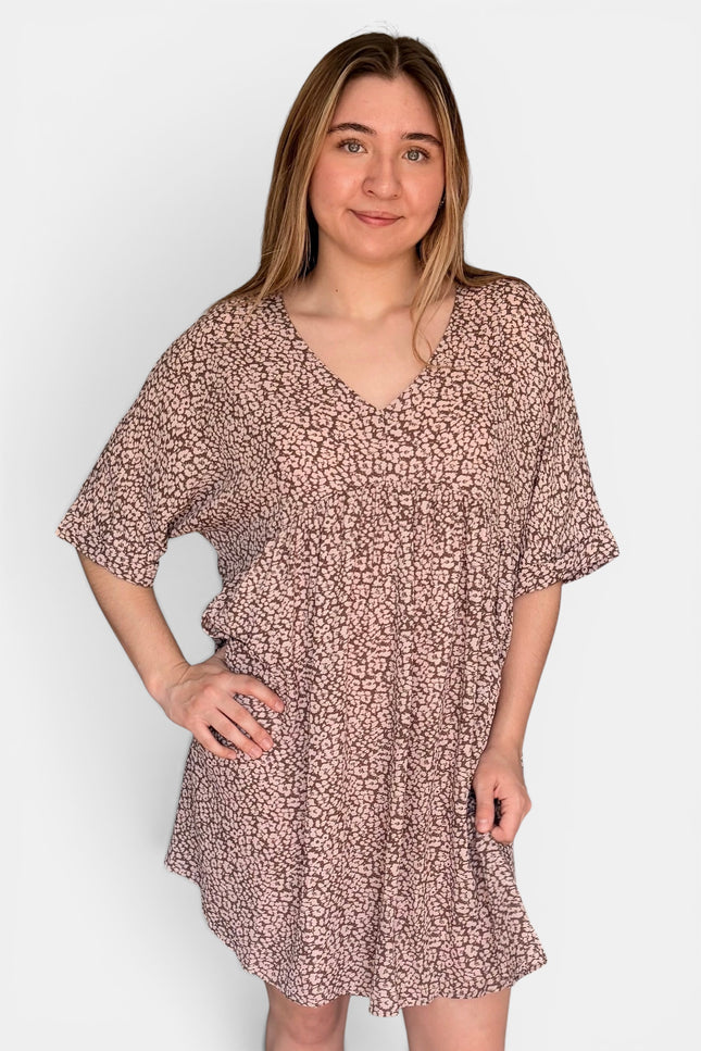 Short-sleeve V-neck dress with a dense small-scale abstract print in dusty pink and brown tones, set against a plain light gray studio background.