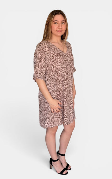 Short-sleeve V-neck dress with a dense small-scale floral or abstract print in muted pink and beige tones, paired with black heeled sandals, against a plain light gray studio background.
