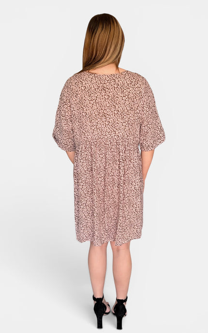 A woman viewed from the back wearing a loose, short-sleeve dress with a dense small floral print in dusty pink and beige, paired with black high heels against a light gray studio background.