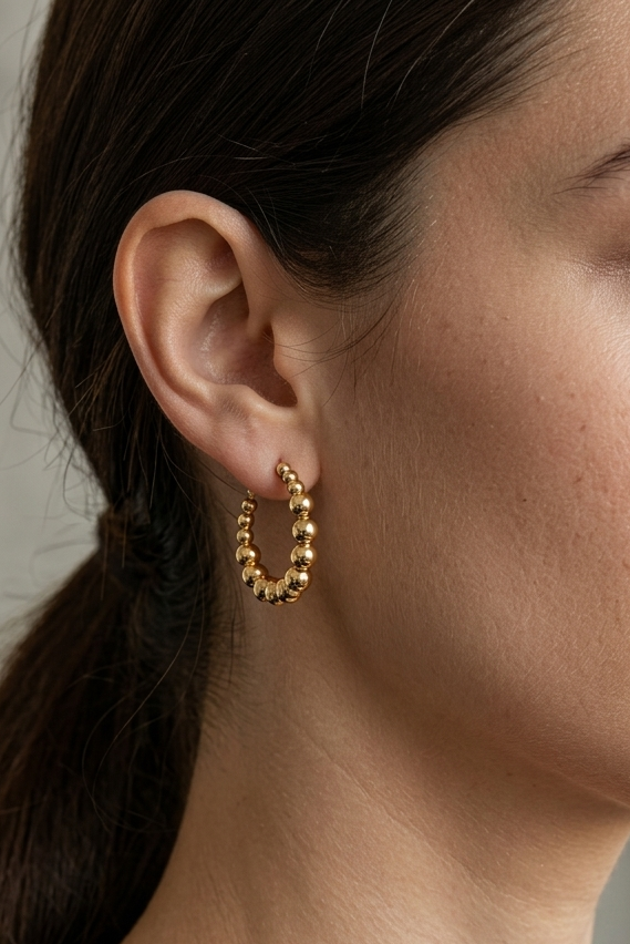 High-detail shot of a woman with dark hair in a low ponytail wearing an 18k gold beaded hoop earring.