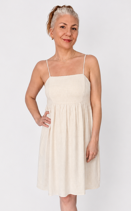A woman modeling a light cream-colored spaghetti-strap mini dress with a gathered waist, paired with white strappy sandals, against a plain light gray background.