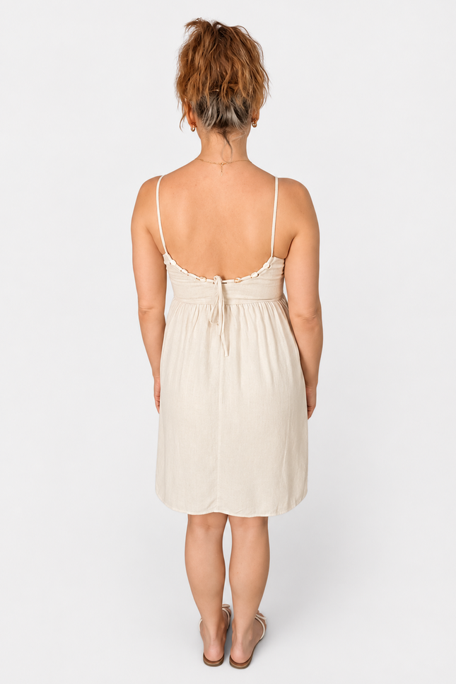 A woman viewed from the back wearing a light cream or off-white spaghetti-strap dress with a tie-back detail, standing against a plain light gray studio background.
