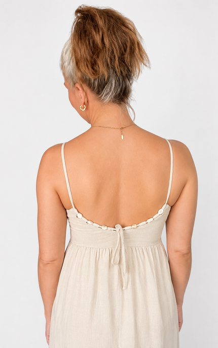 A woman shown from the back wearing a light beige spaghetti-strap dress with a tie-back detail, photographed against a plain light gray studio background.