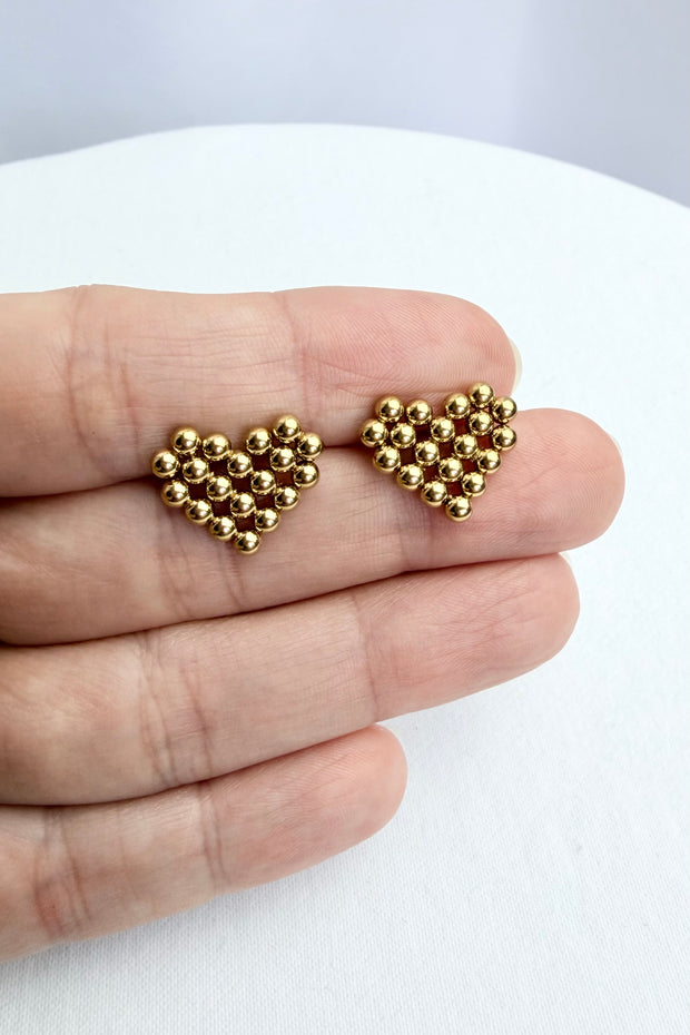 Gold heart-shaped beaded earrings held between fingers on a white background