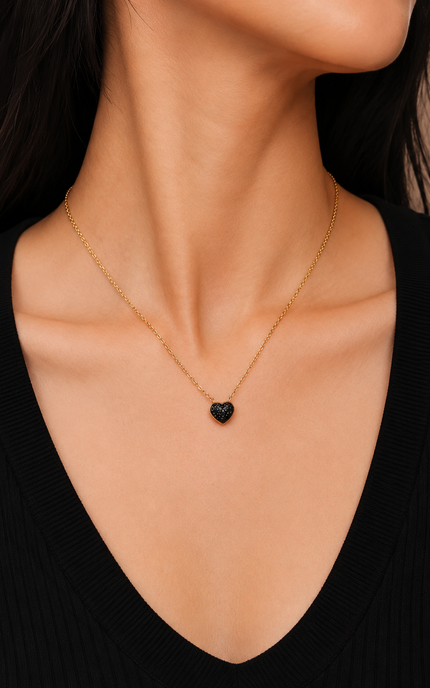 A close-up of a model's neck and chest wearing a delicate gold chain necklace with a small, polished black heart pendant.