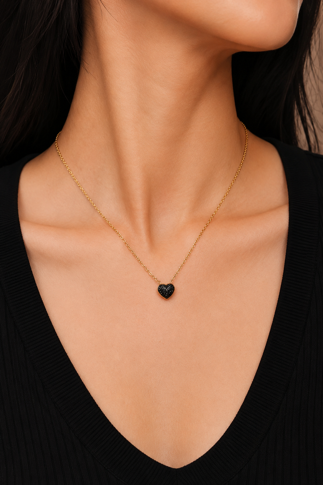 A close-up of a model's neck and chest wearing a delicate gold chain necklace with a small, polished black heart pendant.
