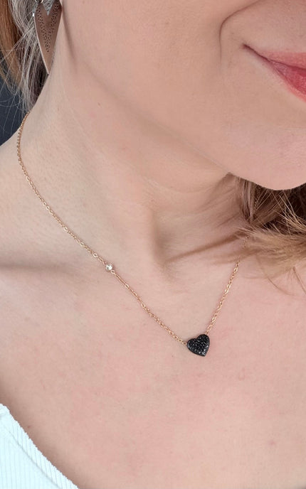 A close-up lifestyle shot of a woman wearing a delicate rose gold chain necklace with a small black heart-shaped pendant and a tiny clear stone accent.