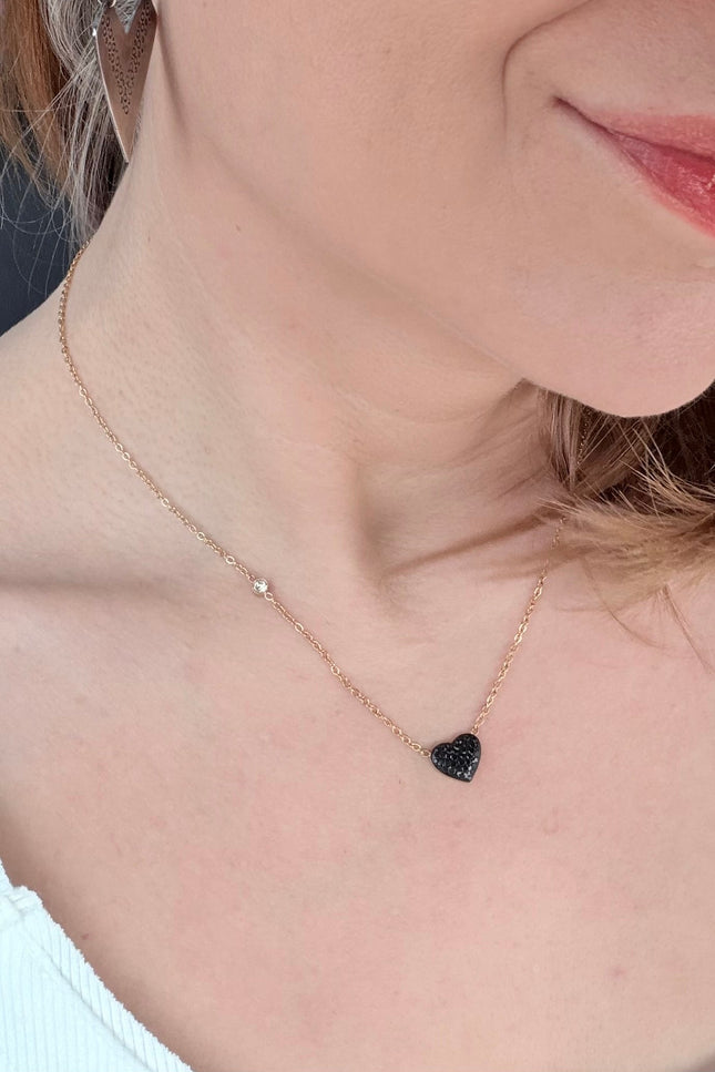 A close-up lifestyle shot of a woman wearing a delicate rose gold chain necklace with a small black heart-shaped pendant and a tiny clear stone accent.