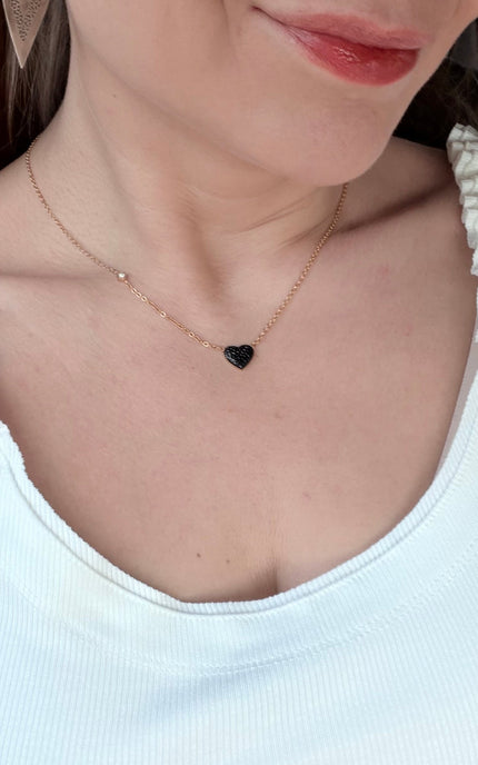 A close-up of a model wearing a delicate gold chain necklace with a small black heart-shaped pendant and a tiny gold bead accent.