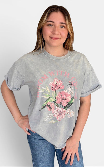 A woman modeling a light gray acid-wash t-shirt with a pink floral graphic and the words 'Bloom with Grace' printed on the front, paired with blue jeans against a light gray background.