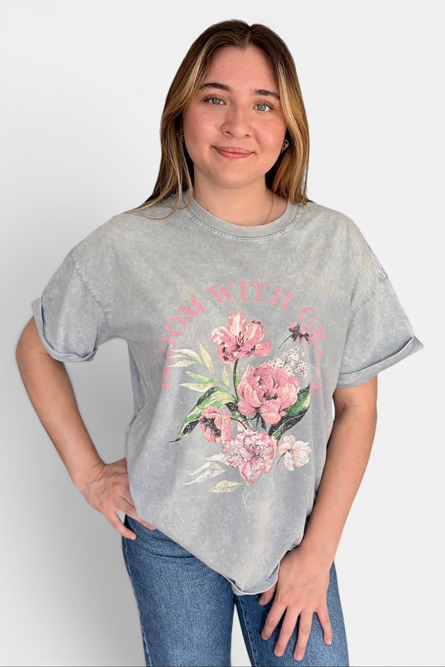 A woman modeling a light gray acid-wash t-shirt with a pink floral graphic and the words 'Bloom with Grace' printed on the front, paired with blue jeans against a light gray background.