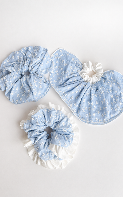 Three light blue fabric hair accessories with a small white floral print, including two scrunchies and one hair clip, arranged on a plain white background.