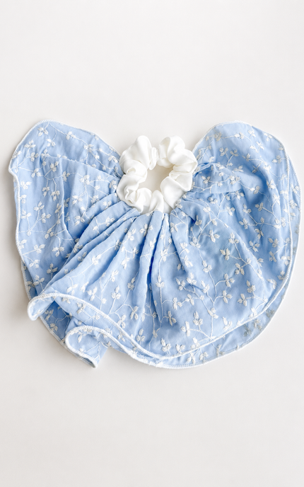 A light blue baby bib with a delicate white floral pattern, gathered around a white fabric scrunchie, laid flat on a light off-white background.
