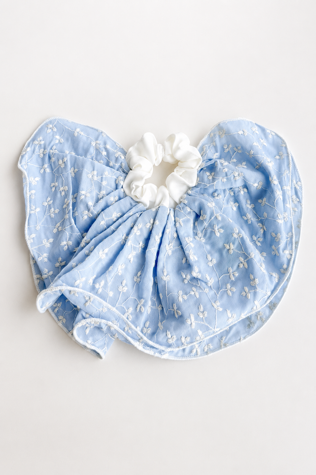 A light blue baby bib with a delicate white floral pattern, gathered around a white fabric scrunchie, laid flat on a light off-white background.