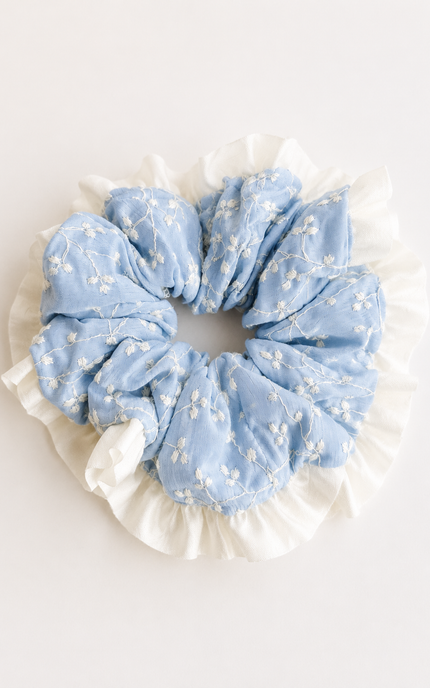 A light blue fabric scrunchie with white floral embroidery and a white ruffled trim, photographed from above on a plain white background.