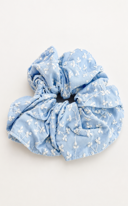 A light blue fabric scrunchie with a delicate white floral embroidery pattern, photographed on a clean, off-white background.