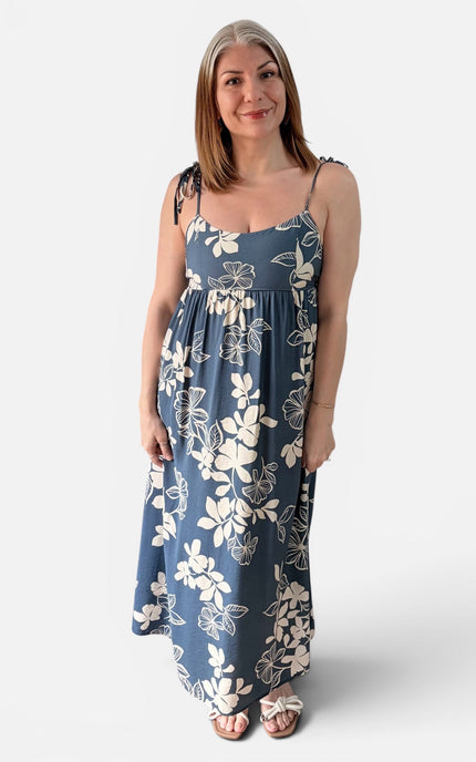 A woman modeling a sleeveless maxi dress with thin straps and a blue floral print, standing against a light gray studio background.