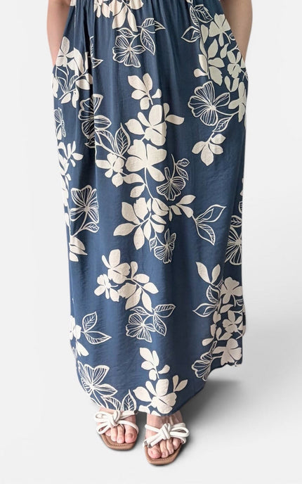 A woman modeling a sleeveless, floor-length maxi dress with a blue base and a cream floral pattern, standing against a plain light gray studio background.