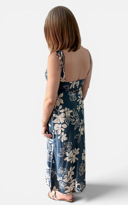 A woman shown from the back wearing a sleeveless maxi dress with a midnight blue and ivory floral pattern, standing against a light gray studio background.