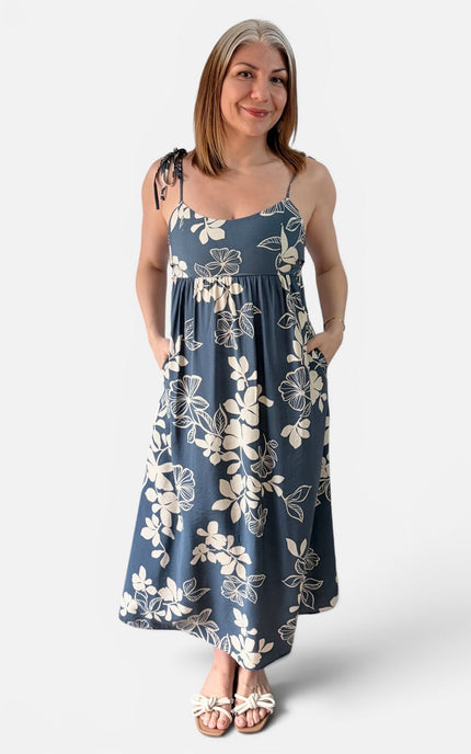 A woman modeling a sleeveless midi dress with a midnight base and ivory floral print, featuring thin straps, side tie details, and side pockets, set against a plain light gray background.