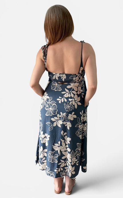 A woman shown from the back wearing a midnight blue floral midi dress with thin straps and a tie detail at the back, photographed against a light gray studio background.