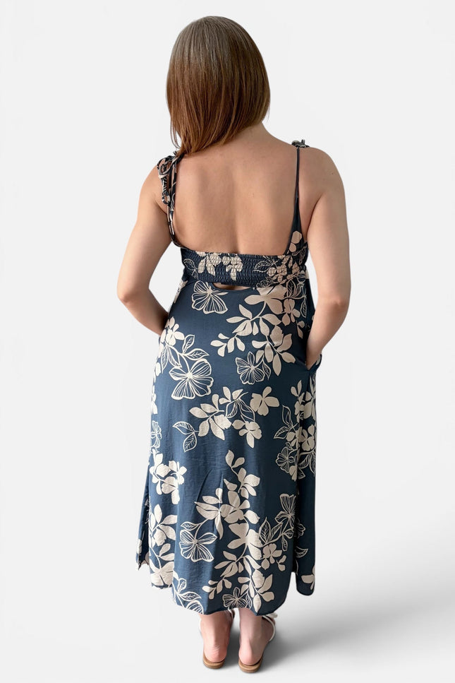 A woman shown from the back wearing a midnight blue floral midi dress with thin straps and a tie detail at the back, photographed against a light gray studio background.