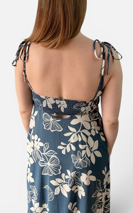 Rear view of a woman wearing a sleeveless midnight blue dress with an ivory floral print and tie-shoulder straps, shown against a light gray studio background.