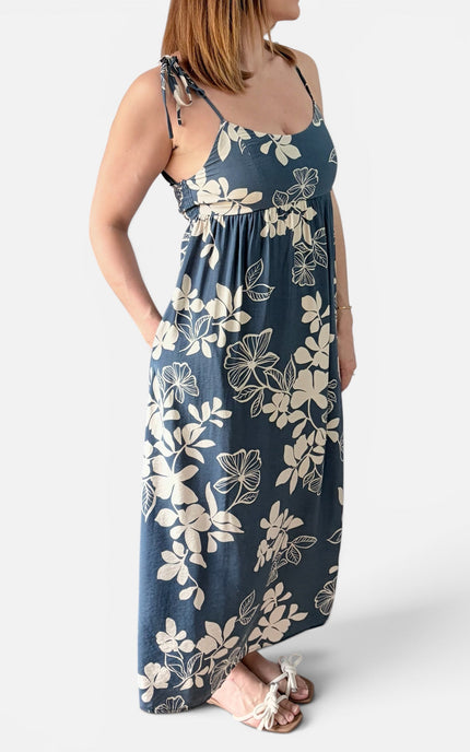 A woman wearing a sleeveless maxi dress with a midnight blue base and an ivory floral print, standing against a light gray studio background.