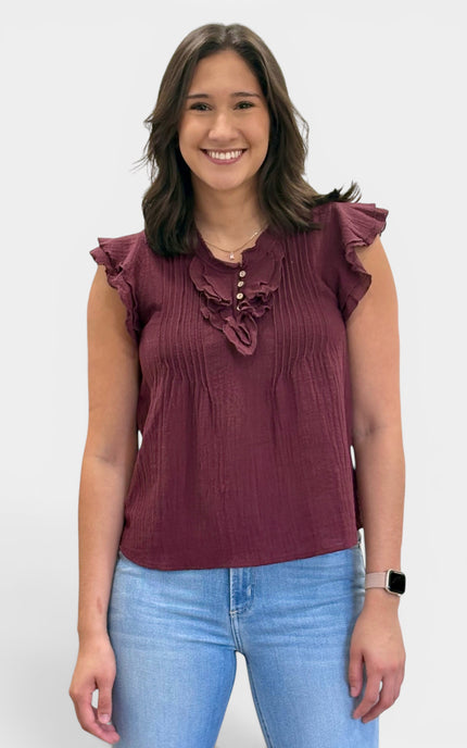 A woman wearing a sleeveless burgundy top with ruffle details on the shoulders and chest, paired with light blue jeans, standing against a plain light gray background.