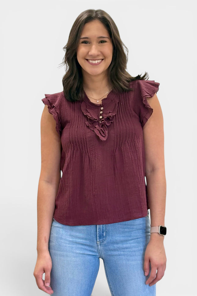 A woman wearing a sleeveless burgundy top with ruffle details on the shoulders and chest, paired with light blue jeans, standing against a plain light gray background.