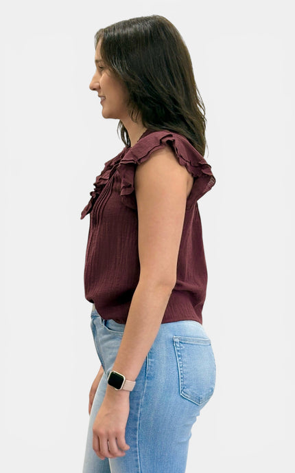 A woman shown in profile wearing a sleeveless ruffled blouse in deep burgundy and light blue jeans, against a plain light gray background.