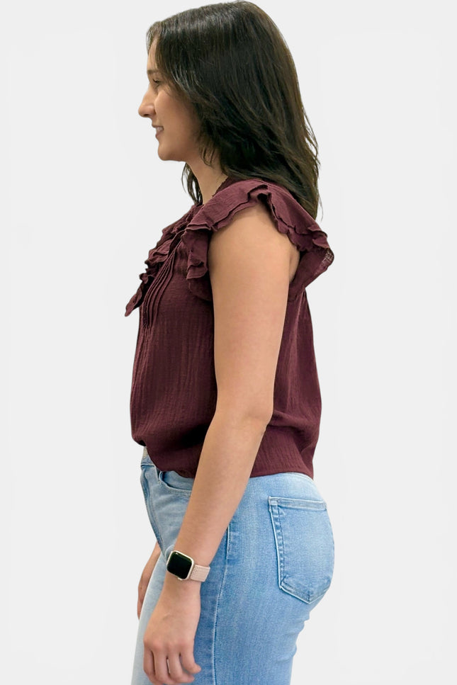 A woman shown in profile wearing a sleeveless ruffled blouse in deep burgundy and light blue jeans, against a plain light gray background.