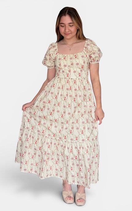 A woman modeling a cream-colored, tiered maxi dress with a small red and green floral print, puff sleeves, and a square neckline against a light gray background.