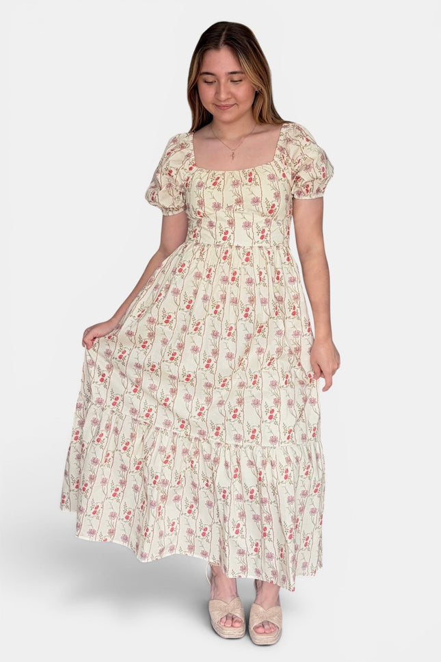 A woman modeling a cream-colored, tiered maxi dress with a small red and green floral print, puff sleeves, and a square neckline against a light gray background.