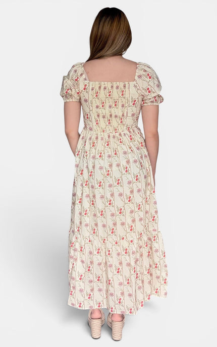 A woman viewed from the back wearing a cream midi dress with a red and green floral print, puff sleeves, and a smocked bodice, paired with beige strappy heels against a light gray studio background.