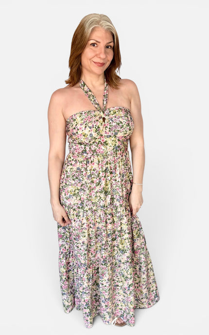 A woman modeling a strapless halter-neck maxi dress with a dense multicolor floral print on a light cream background, set against a plain light gray studio backdrop.