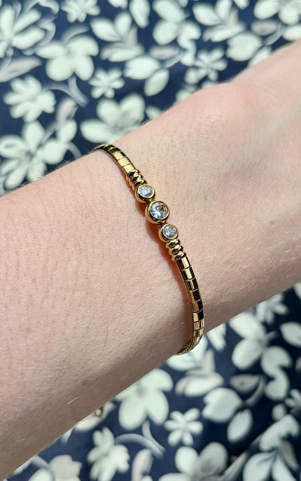A gold bangle bracelet with three clear round gemstones, shown worn on a person's wrist against a navy and white floral fabric background.
