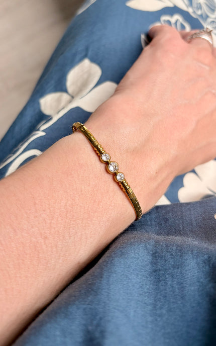 A close-up of a person's wrist wearing a textured gold bracelet with three small clear gemstones, resting on blue floral fabric.