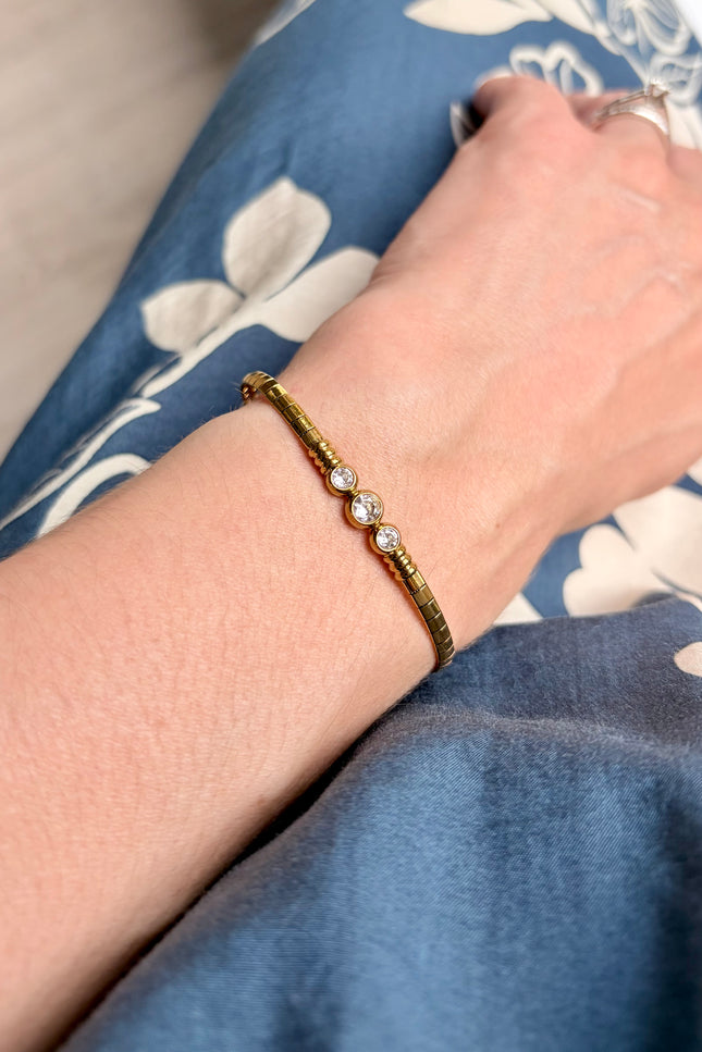 A close-up of a person's wrist wearing a textured gold bracelet with three small clear gemstones, resting on blue floral fabric.